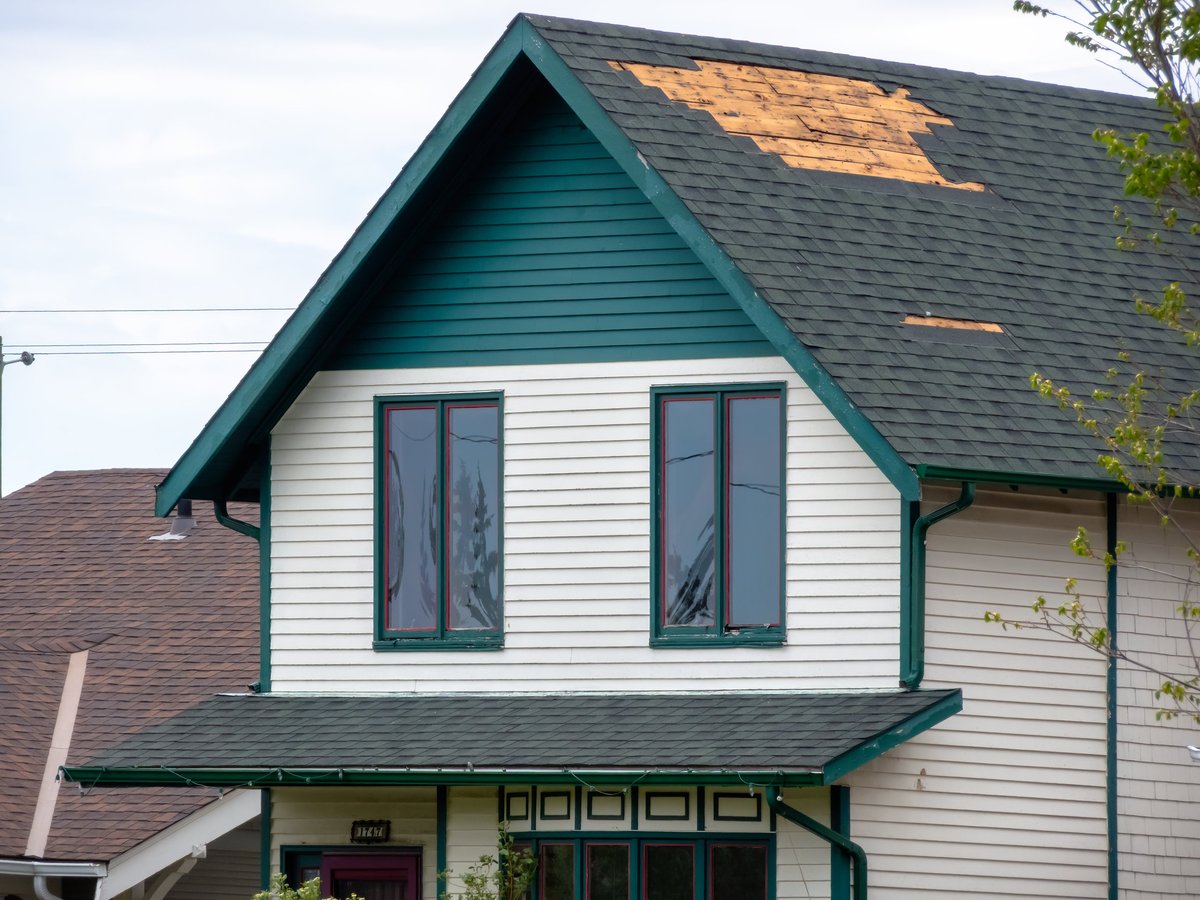 Weathered residential house exterior with a damaged roof, missing shingles and exposed wood